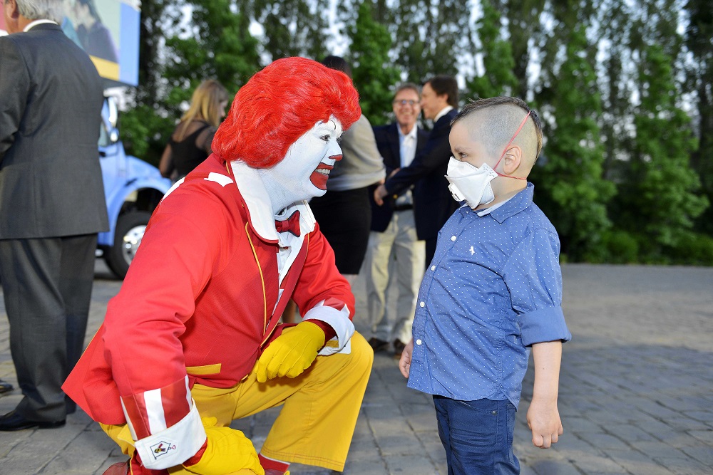 Ronald McDonald se arrodilla para quedar al nivel de un niño que lleva un tapabocas puesto. Detrás se ven varias personas sonriendo.